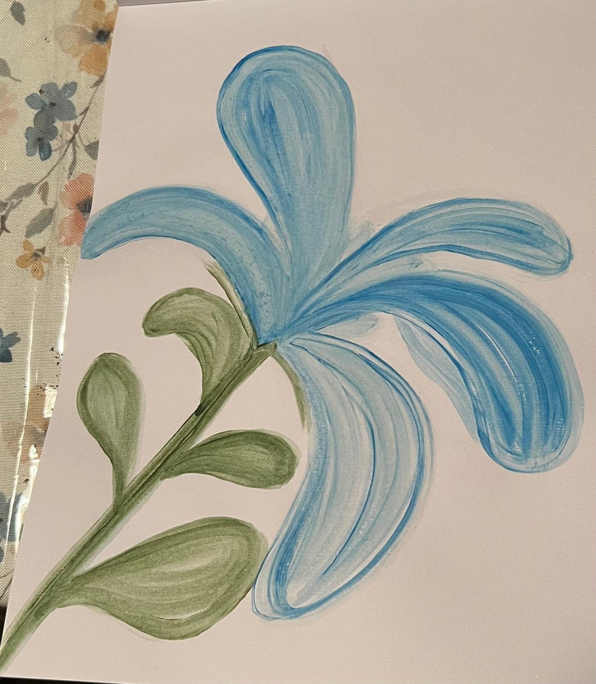 A single blue flower with long flowing petals and green leaves, photographed on paper.